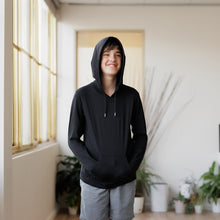 Load image into Gallery viewer, Teenage boy standing in a room wearing a Sensory Friendly Clothing long sleeved hooded t-shirt with the hood up over his head.  He has his hands in the kangaroo pocket and looks relaxed and content.