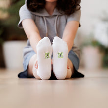 Load image into Gallery viewer, Child sitting on the ground with legs outstretched wearing a pair of comfy white Sensory Friendly Clothing Seamless Feel Socks.  The non irritating printed black and green Sensory Friendly Clothing logo is shown on the soles of the socks.
