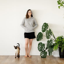 Load image into Gallery viewer, Teenage girl standing against a white wall wearing a grey Sensory Friendly Clothing long sleeved hooded t-shirt and black shorts.  The hood is over her head and her hands are inside the kangaroo pouch pocket.  Looks relaxed and comfortable in her clothing.