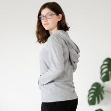 Load image into Gallery viewer, Teenage girl standing sideways to the camera wearing the Sensory Friendly Clothing grey long sleeved hooded t-shirt with the hood down.  She has her hands in the kangaroo pouch pockets and she looks relaxed and calm.