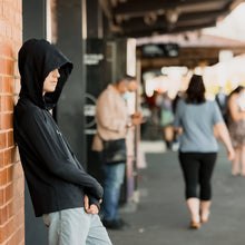 Load image into Gallery viewer, Boy learning against a brick wall at a busy railway station.  He is wearing a black Sensory Friendly Clothing long sleeved hooded t-shirt with the hood over his head so that only part of his face is visible.  He has his thumbs through the thumb holes of the sleeved with his arms crossed over each other relaxed against his torso.  He looks comfortable and relaxed in a busy environment.