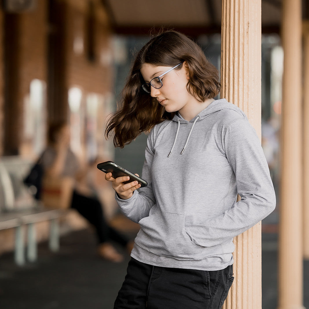 Girl looking down at her phone while learning against a poll in a noisy, busy location.  She is wearing a grey merle Sensory Friendly Clothing hooded long sleeved t-shirt and black shorts.  She has her hand in the kangaroo pouch pocket and looks relaxed.