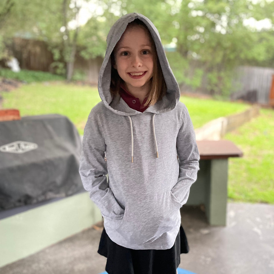 Young girl wearing a grey merle Sensory Friendly Clothing hooded long sleeved t-shirt.  She has the hood comfortable up over her head and her hands in the kangaroo pouch pockets.  She is smiling and content.