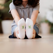 Load image into Gallery viewer, Child sitting on the ground with legs outstretched wearing a pair of comfy white Sensory Friendly Clothing Seamless Feel Socks.  The non irritating printed black and green Sensory Friendly Clothing logo is shown on the soles of the socks.  
