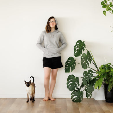 Teenage girl standing against a white wall wearing a grey Sensory Friendly Clothing long sleeved hooded t-shirt and black shorts.  The hood is over her head and her hands are inside the kangaroo pouch pocket.  Looks relaxed and comfortable in her clothing.