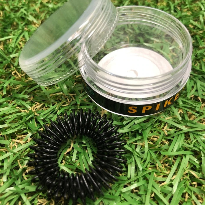 A black Kaiko Spikey Fidget placed on the grass with it's clear plastic container sitting next to it with the lid open.